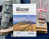 Przewodnik "Ruszaj w Bieszczady" z widokiem na góry i turystów. Obok rzeźba z drewna i świeca z napisem "all you need is in your Home".