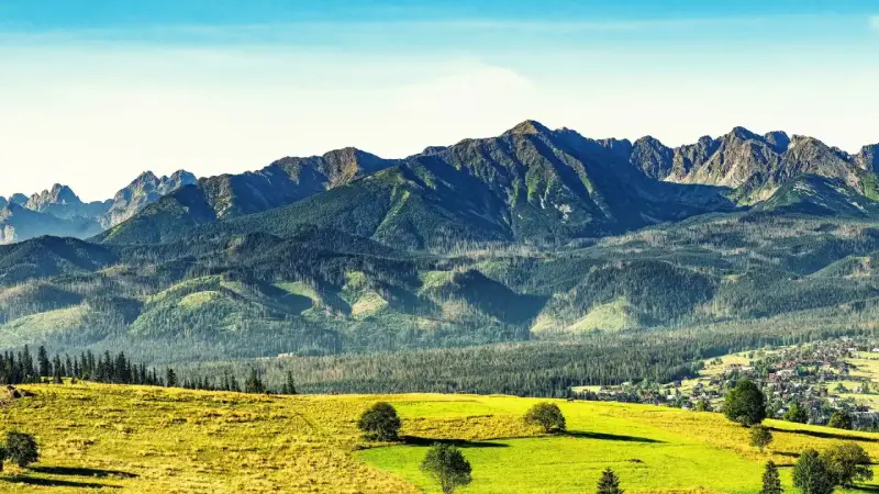 Widok na Tatry po angielsku: zielone wzgórza, lasy i skaliste szczyty. W oddali widać małą wioskę.